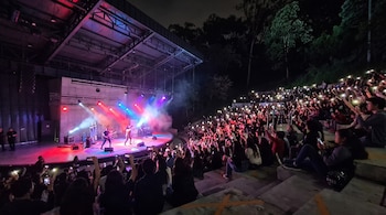 Un concierto nocturno en un anfiteatro al aire libre con público sosteniendo teléfonos iluminados, una banda en el escenario, instrumentos y luces de colores.