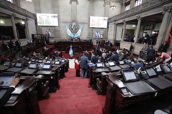 Interior de un hemiciclo parlamentario con bancadas de madera, pantallas individuales, alfombra roja, y legisladores. Al fondo, pantallas grandes y banderas nacionales