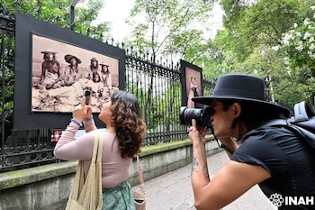 Dos personas, una con teléfono y otra con cámara, fotografían grandes imágenes históricas en blanco y negro montadas en una valla al aire libre