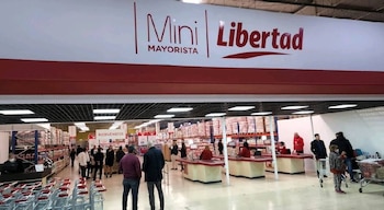 Entrada de un supermercado con el cartel Mini Libertad Mayorista, cajas registradoras, carritos de compra, clientes y estanterías con productos