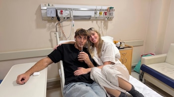 Un hombre y una mujer sentados en una cama de hospital. Detrás hay equipo médico y un mueble. El hombre bebe con pajita y la mujer sonríe.