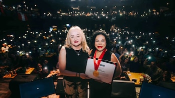 Dos mujeres de pie en un escenario. Una sostiene un certificado y una medalla. Detrás, el público en la oscuridad sostiene teléfonos encendidos