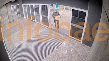 Un hombre con abrigo con capucha camina por un pasillo con puertas de vidrio mientras sostiene una caja de cartón y un teléfono celular