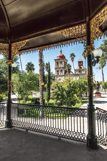 Vista do quiosque com janela para a praça com árvores, palmeiras, igreja com piscina vermelha e torre do relógio