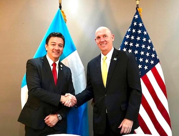 Dos hombres vestidos con traje y corbata se dan la mano, con la bandera de Guatemala y la bandera de Estados Unidos detrás de ellos