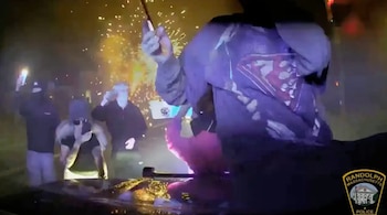 Escena nocturna oscura con gente reunida; un individuo en primer plano manipula un objeto con chispas, mientras otros observan y graban con teléfonos