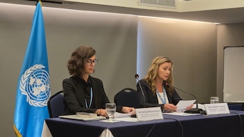Dos mujeres sentadas frente a una mesa con micrófonos, credenciales de identificación, papeles y la bandera de las Naciones Unidas