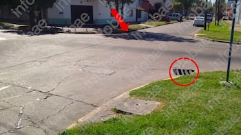 Esquina de calle con pavimento de asfalto agrietado, un bordillo, acera y un área de césped. Hay una rejilla de desagüe abierta en el césped