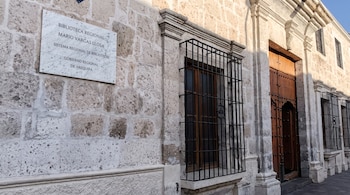 Fachada de piedra clara con una placa de mármol, una ventana con rejas de hierro negro y una gran puerta de madera.