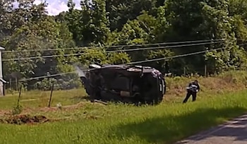 Vista aérea de una SUV negra volcada a un lado de una carretera rural con humo saliendo. Un hombre corre a la derecha, cerca de un área de árboles y vegetación