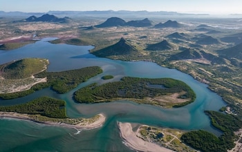 Vista aérea de una bahía azul con islas de manglares verdes, playas de arena y montañas en el horizonte bajo un cielo despejado