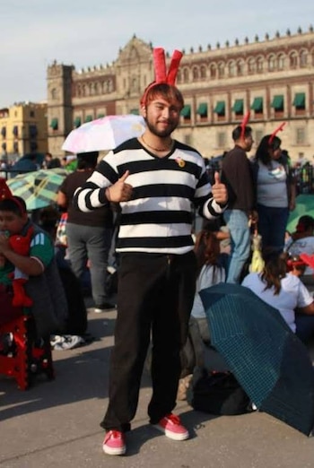 Jovem de suéter colorido, calça preta e sapatos rosa, sorrindo e mostrando os polegares para cima, usando bandana vermelha. Ao fundo, pessoas e edifícios antigos