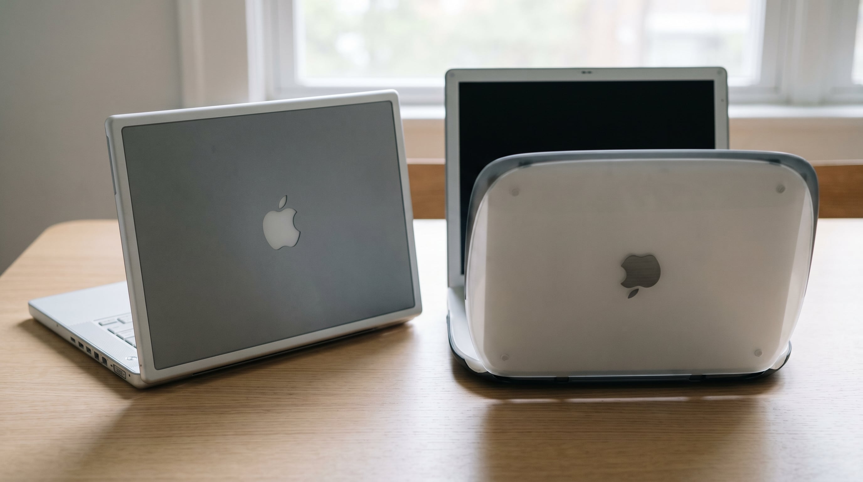 Dos portátiles Apple de diseño clásico, inspirados en PowerBook e iBook, se exhiben en una mesa de madera clara, con uno cerrado y otro abierto, destacando sus logos de la manzana. (Imagen Ilustrativa Infobae)