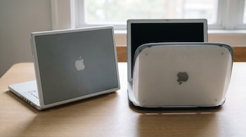 Dos portátiles Apple plateados y blancos, uno cerrado a la izquierda y otro abierto a la derecha, sobre una mesa de madera clara con una ventana al fondo.