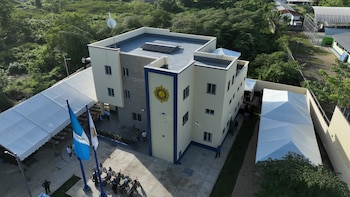 Edificio de varios pisos crema y azul con emblema circular, dos banderas, motocicletas estacionadas, personas, dos carpas blancas y árboles