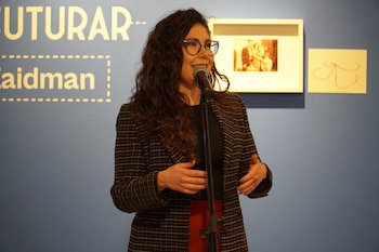 Mujer de cabello rizado con gafas habla por micrófono, frente a una pared azul con texto en dorado y dos marcos, uno con una foto antigua