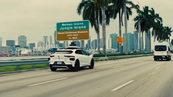 Un coche autónomo Waymo blanco circula por una autopista con palmeras, el horizonte de Miami, una señal de tráfico y un furgón blanco delante