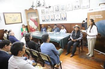 Personas reunidas en una sala con piso de madera, retratos en la pared y una mujer de pie junto a un panel informativo mientras habla al grupo