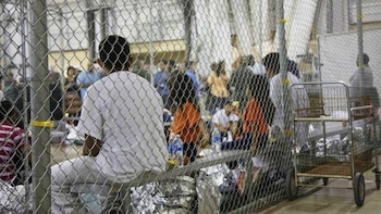 Personas, incluyendo niños, vistas desde atrás, detrás de una valla de tela metálica en una instalación interior. Mantas y botellas de agua son visibles