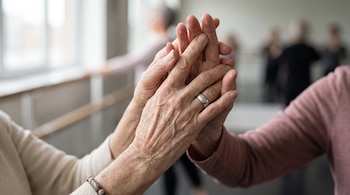 Primer plano de dos manos de adultos mayores entrelazadas; una mano lleva un anillo y una pulsera. Fondo difuminado de una sala de danza con personas.