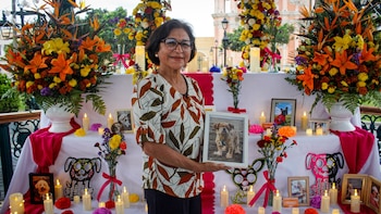 Una mujer con gafas sostiene un portarretratos frente a una mesa decorada con flores, velas y fotografías de animales domésticos