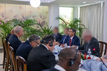 John Ratcliffe, con traje azul y corbata roja, sentado en una mesa de conferencias con otras personas, algunas con micrófonos, en una sala con plantas