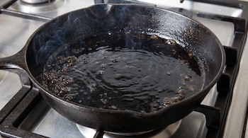 Una sartén de hierro fundido negra con aceite oscuro burbujeando y humo ligero sobre una estufa de acero inoxidable.