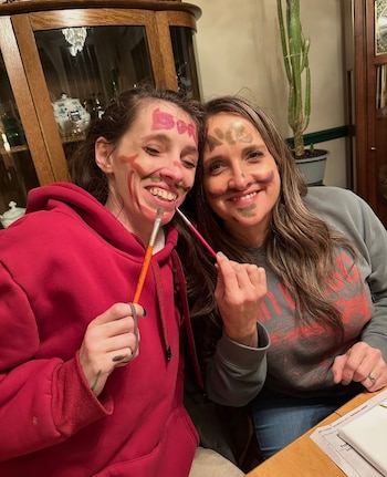 Dos mujeres sonrientes con pintura multicolor en sus rostros, sosteniendo pinceles. La mujer de la izquierda lleva una sudadera roja, la de la derecha una gris