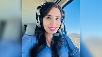 Una mujer de cabello oscuro y blusa azul lleva auriculares grandes con micrófono dentro de la cabina de una aeronave junto a una ventana