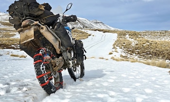 Rueda trasera de una motocicleta con cinchas rojas para nieve sobre un camino cubierto de nieve frente a colinas montañosas