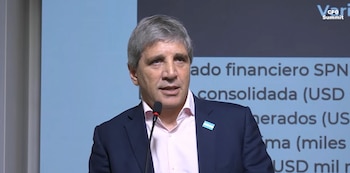 Un hombre con saco oscuro, camisa clara y un broche de la bandera de Argentina habla ante un micrófono delante de una pantalla