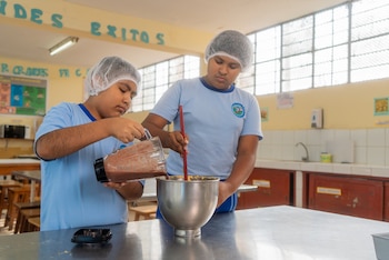 Dos escolares con cofias para el cabello vierten una mezcla de licuadora en un tazón metálico sobre una mesa de cocina escolar