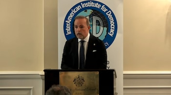 Hombre con barba y traje oscuro en podio con placa dorada. Detrás, bandera del InterAmerican Institute for Democracy con un globo terráqueo