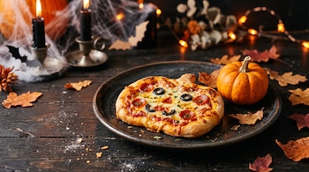 Mini pizza con forma de calabaza en un plato negro junto a una calabaza pequeña, hojas secas, velas y telarañas sobre una mesa de madera oscura.