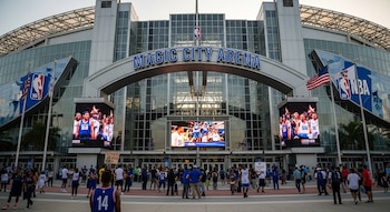 Vista frontal de la entrada del Magic City Arena con su nombre en letras azules, grandes pantallas mostrando jugadores y personas afuera.
