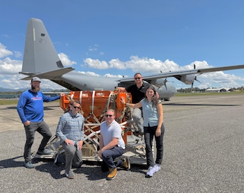 Cinco personas posan junto a una caja naranja asegurada con correas sobre un palé, frente a un avión militar gris en una pista