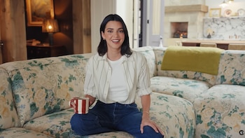 Kendall Jenner sentada sonriendo en un sofá floral con una manta verde, sosteniendo una taza. Al fondo se ve una chimenea y detalles de diseño interior