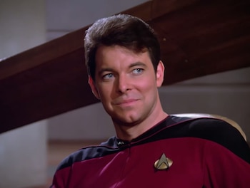 Primer plano de Jonathan Frakes con el cabello castaño y ojos azules, vistiendo un uniforme rojo y negro de Starfleet con una insignia dorada en el pecho