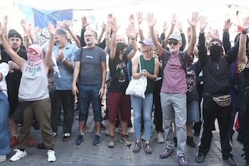 Un grupo de personas en una plaza de adoquines levanta las manos abiertas hacia arriba frente a pancartas y carpas al fondo