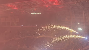Interior de un estadio con iluminación roja y humo. Se observan dos chorros de chispas en el centro, una multitud en las gradas y carteles de BMO Stadium y Ford