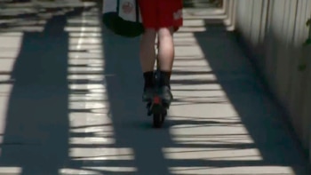 Parte trasera de una persona en patinete eléctrico sobre pavimento. Viste pantalones cortos rojos, lleva una mochila y hay sombras de barandales en el suelo