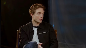 Primer plano de Robert Pattinson, un hombre de cabello castaño, sentado y mirando a la derecha, vistiendo una chaqueta negra de cuero sobre una camiseta blanca
