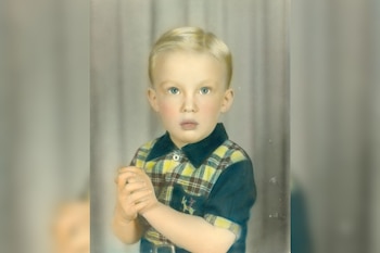 Fotografía de un niño rubio de ojos claros con camisa a cuadros y las manos entrelazadas frente a un telón