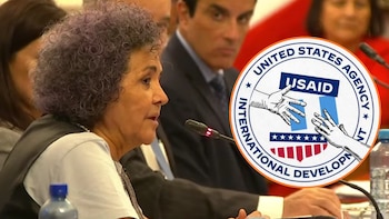 Una mujer de cabello rizado habla ante un micrófono en una mesa con otras personas, junto al logotipo circular de USAID superpuesto