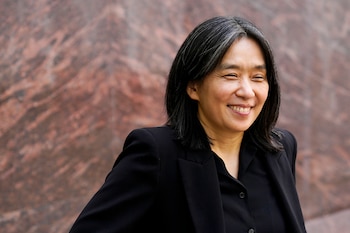Retrato de una mujer con cabello oscuro y canas sonriendo con los ojos cerrados, vestida con un blazer y camisa negra, frente a una pared texturizada marrón