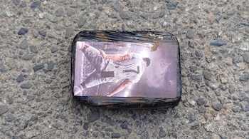 Un bulto rectangular envuelto en plástico negro con una fotografía de un futbolista con el número 7 en su espalda. El bulto yace sobre asfalto y gravilla.