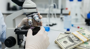 Manos enguantadas manipulan un microscopio óptico que sostiene un portaobjetos con muestras; al lado, fajos de billetes de cien dólares sobre una mesa de laboratorio.