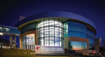 Fachada exterior del Centro Global Davis de UNMC por la noche, con grandes ventanales curvos y una pasarela elevada, iluminada bajo un cielo azul oscuro