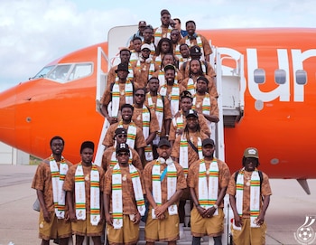 Mais de 20 membros da equipe de Gana estão nos degraus e em frente ao avião laranja. Eles usam uniformes marrons e lenços brancos com as cores de Gana