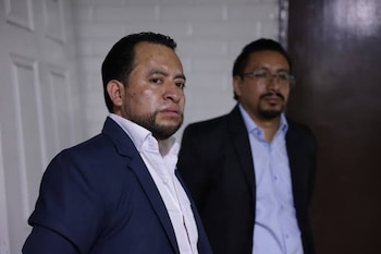 Primer plano de un hombre con traje azul y camisa blanca, con barba. Detrás, otro hombre con traje oscuro, camisa azul y gafas